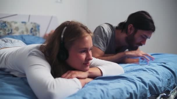 Женщина слушает музыку в наушниках и мужчина с помощью планшета в постели — стоковое видео