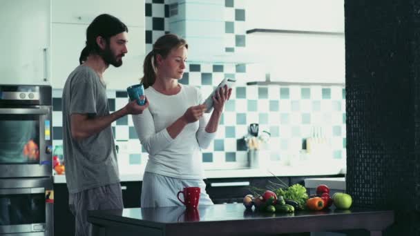 Mujer usando una tableta de pie en la cocina con el hombre sosteniendo una taza — Vídeo de stock