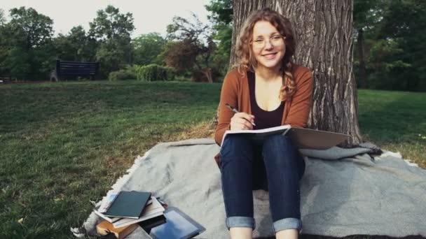 Portret van een glimlachende vrouwelijke student zitten onder de boom in het park — Stockvideo