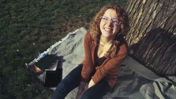 Портрет улыбающейся студентки, сидящей под деревом в парке — стоковое видео