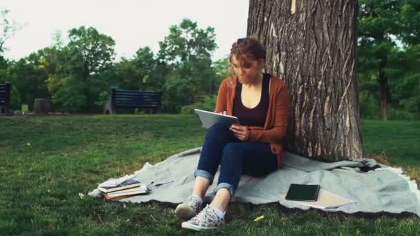 Étudiante assise sous un arbre dans le parc et utilisant une tablette — Video