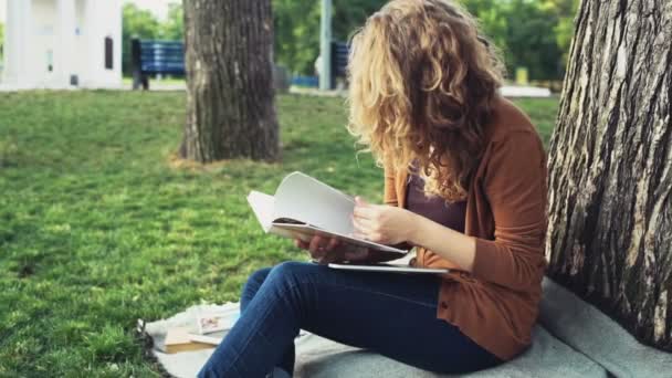 Kvinnlig student vända anteckningsboken under ett träd i parken slow motion — Stockvideo