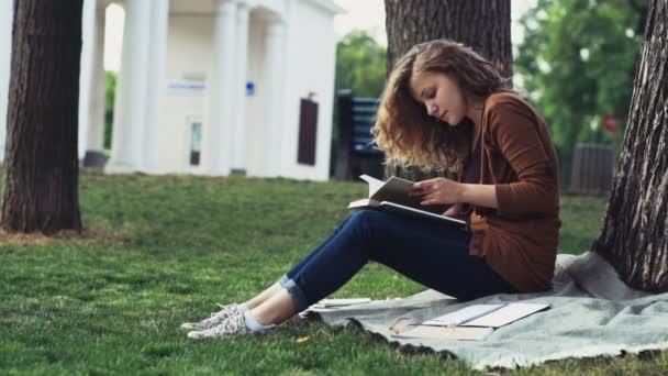 Estudante do sexo feminino lendo sob a árvore no parque câmera lenta — Vídeo de Stock