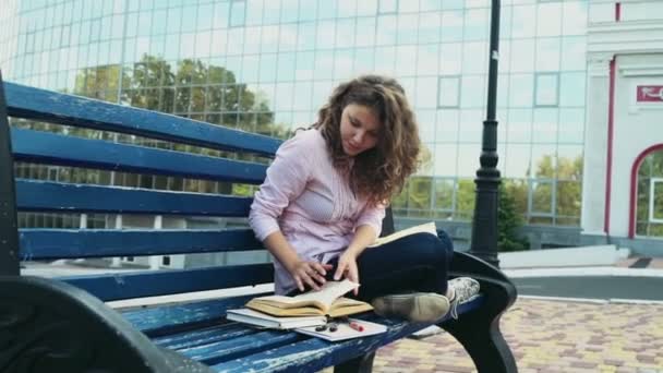 Estudante feminino sentado em um banco perto do edifício moderno câmera lenta — Vídeo de Stock