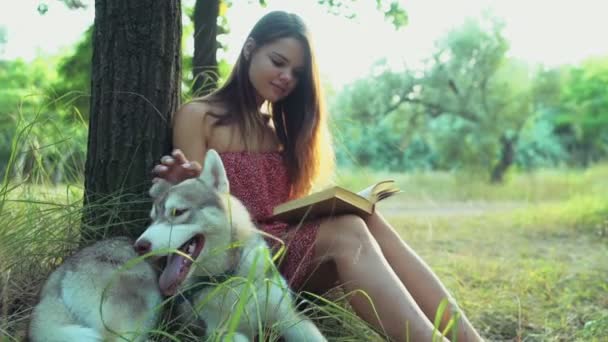 Tonårsflicka läser en bok och smeker en husky hund i skogen slow motion — Stockvideo