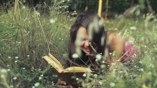 Riéndose adolescente chica con un libro pone en un campo hierba cámara lenta — Vídeo de stock