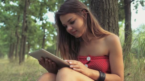 Mujer joven se sienta junto a un árbol y utiliza una tableta de cámara lenta — Vídeos de Stock