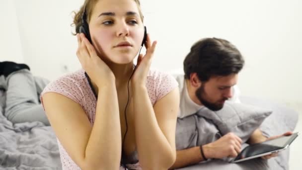 Mujer escuchando música en auriculares, hombre usando tableta en la cama — Vídeo de stock