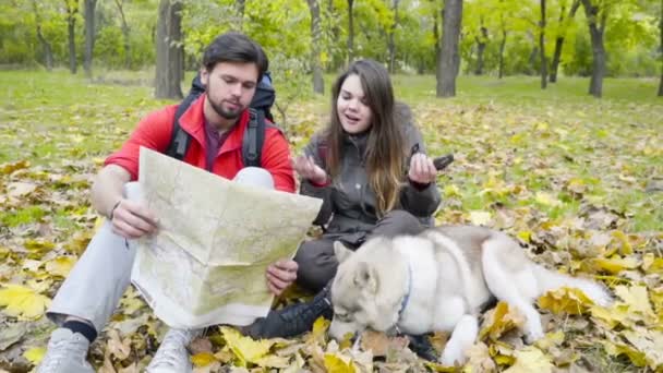 Pareja de excursionistas y un perro husky en el bosque de otoño utilizando un mapa de papel — Vídeos de Stock