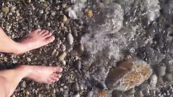 海水が流れる海岸の小石に足を踏み入れた男の姿です。垂直ビデオ — ストック動画