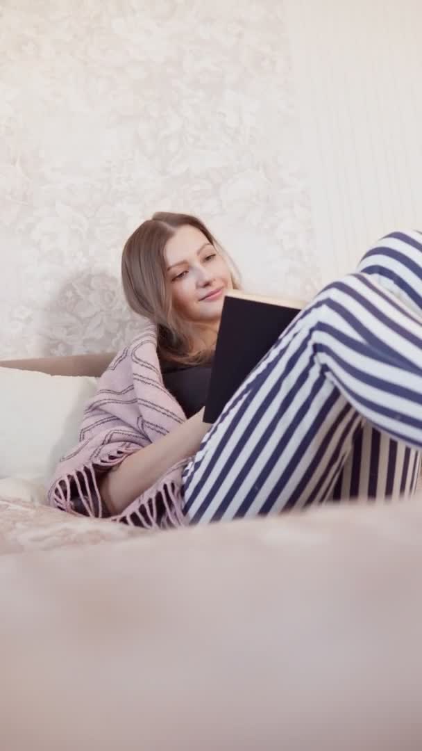 Молода дівчина читає книжку. домашній одяг лежить у ліжку. Відпочиньте розслабитися від хорошого настрою концепції способу життя. вертикальне відео — стокове відео