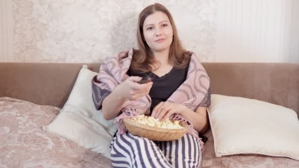 Молодая девушка с длинными рыжими волосами сидит на кровати и смотрит телевизор и ест попкорн. оставаться дома во время карантина — стоковое видео