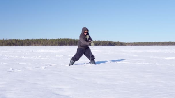 Jovem pescador broca gelo com uma broca de gelo em um lago de inverno. Pesca no gelo — Vídeo de Stock