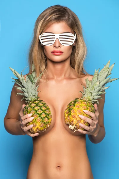 Young sexy woman happy smiling only in glasses posing on blue background with pineapple who cover her breast — ストック写真
