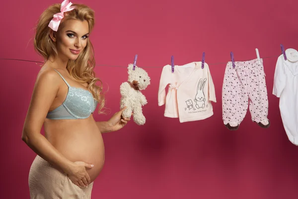 Беременная женщина позирует рядом с детской одеждой на розовом фоне — стоковое фото
