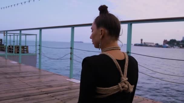 一个身穿黑色紧身衣 头发整整齐齐地堆在一起的女孩 端坐在海边的一个码头上 一个阳光灿烂的早晨 被绳子捆着的躯体 — 图库视频影像