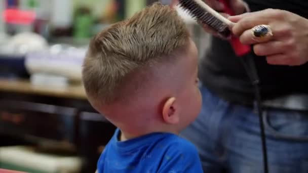 Un styliste portant un masque covid-19 fait une coiffure élégante. Une coupe de cheveux d'enfant dans le salon, un petit garçon se fait couper les cheveux dans le salon de coiffure. Stylisme pour enfants. — Video