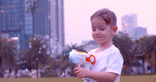 A preschool boy in a city park blows bubbles from a pistol in the park. The kid is having fun with the aunts, the kids are running. — Stock Video