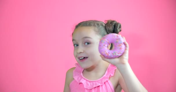 Привлекательная маленькая девочка держит пончики с глазурью на руках улыбаясь смотрит в камеру и приносит их к ее лицу глядя на коллекцию сладких пончиков с розовой глазурью. Портрет ребенка — стоковое видео