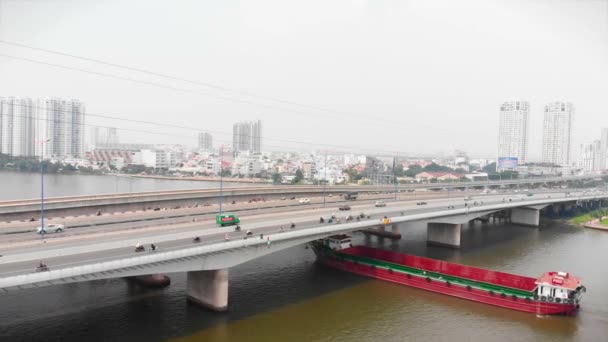 Tiro voo drone aéreo com vista para o rio com navios porta-contentores e contra o pano de fundo de belas grandes edifícios da cidade com carros e motocicletas passando. Megápolis, Ho Chi Minh — Vídeo de Stock