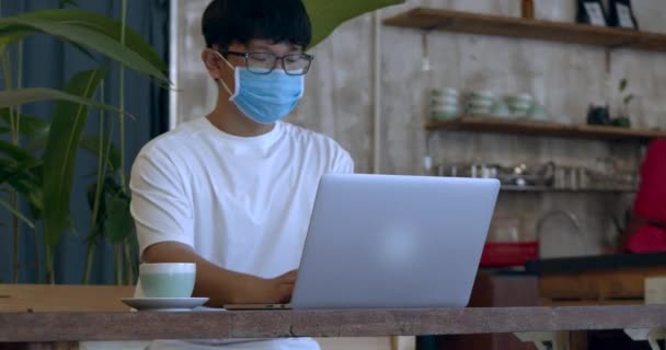 Een jongeman met een bril. man tiener in beschermende masker, student werken op laptop van cafe en het drinken van koffie. — Stockvideo