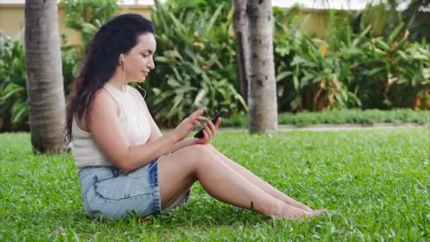 European Woman användning med mobiltelefon lyssna musik via hörlurar på naturen koppla av i parken sitter på gräset eller gräsmatta på gatan. — Stockvideo