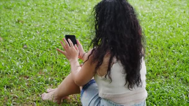 Ευρωπαϊκή γυναίκα κάνει χρήση ενός tablet ή τηλεφώνου με ένα smartphone, φύλλα ή εκτυπώσεις με το δάχτυλό της, επικοινωνεί χρησιμοποιώντας την εφαρμογή, χαλαρώνοντας στο δρόμο, ενώ κάθεται στο γρασίδι στο πάρκο. — Αρχείο Βίντεο
