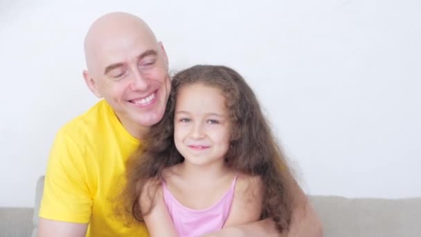 Famille heureuse, papa aimant avec une petite fille sont assis à la maison sur le canapé en regardant la caméra, père donne câlins et un baiser à sa fille mignonne bien-aimée, enfant se réjouit dans les soins des papas d'elle. — Video