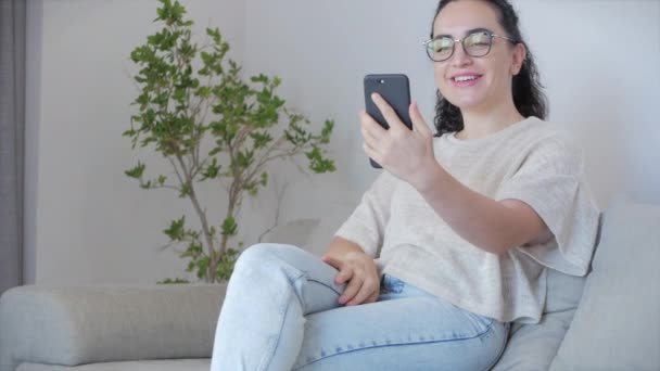 Счастливая молодая европейская расслабляющая женщина или девушка, глядя в телефон, делает селфи сидя дома на диване, концепция видеочата vlogger. — стоковое видео