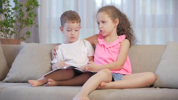 Enfants mignons, sœur avec un frère cadet, regardant un livre magazine manuel dictionnaire tablette lire des contes de fées, les enfants lisent des livres assis sur le canapé, préscolaire et l'éducation scolaire. — Video