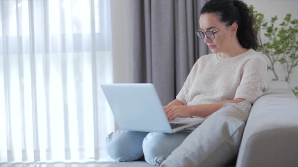 Młoda poważna kobieta Pracując na laptopie, siedząc na kanapie w domu, businesswoman siedzi w domu prace zdalnie na laptopie lub smartphone sprawdza pocztę do nauki pracy online w domu. — Wideo stockowe