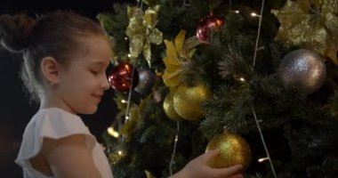 Çocuk, Noel ışıkları ve kırmızı ve sarı toplarla Noel ağacına asılan Noel süslerine seviniyor. Noel ağacını topla süslemek. 4K