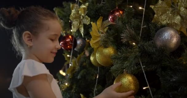 El niño se regocija por las decoraciones navideñas que se cuelgan en el árbol festivo con luces de Navidad y bolas rojas y amarillas. Decorar un árbol de Navidad con una pelota. 4K — Vídeos de Stock