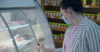 Coronavirüs salgınından maske takmış genç bir kadın bir süpermarketin bakkal bölümünde insanların yiyecek satın aldığı konsepti satın alıyor..