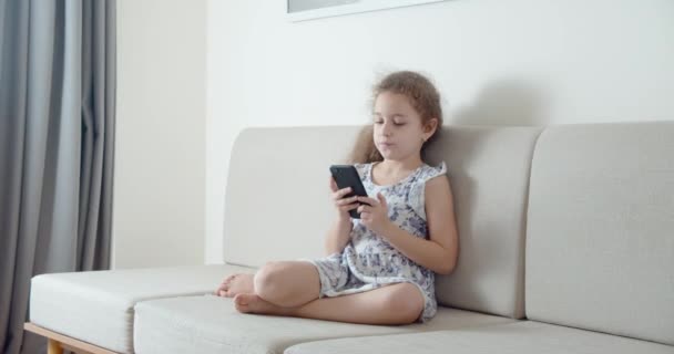 Glückliches kleines Mädchen oder Kind, das zu Hause entspannt mit einem Smartphone kuschelt, auf dem Sofa sitzt Tochter und Sohn, auf den Bildschirm eines Mobiltelefons schaut, Zeichentrickfilme anschaut. — Stockvideo