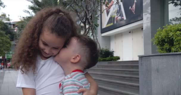 Crianças, uma irmãzinha encaracolada está a abraçar o irmão. Irmãozinho e irmã abraçam e beijam na frente da câmera. Família, duas crianças na rua fazendo pose para a câmera. — Vídeo de Stock