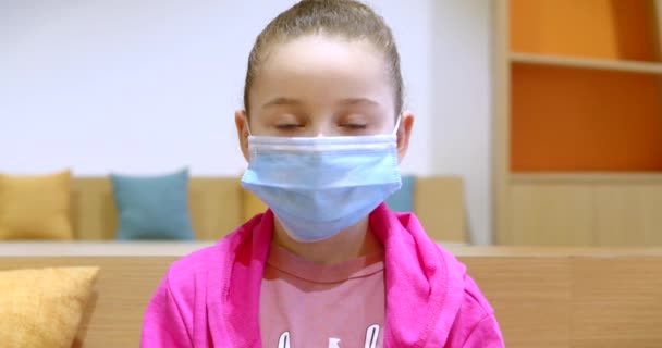 Portretul unei fetițe într-o mască de protecție copilul se uită la camera care stă pe canapea acasă, copil într-o mască medicală. Pandemie, covid - 19. Conceptul de pandemie din copilărie . — Videoclip de stoc