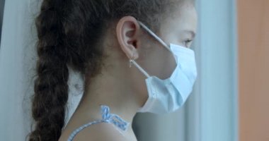 Koruyucu maske takmış şirin bir çocuk ya da küçük bir kızın portresi üzgün bir hayal kırıklığıyla pencereden dışarı bakıyor. Tıbbi maskeli bir kız. Salgın, covid - 19.