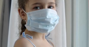Koruyucu maske takmış şirin bir çocuk ya da küçük bir kızın portresi üzgün bir hayal kırıklığıyla pencereden dışarı bakıyor. Tıbbi maskeli bir kız. Salgın, covid - 19.