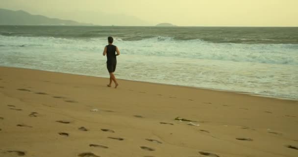 Jovem caucasin homem estão se exercitando com ao ar livre correndo com um belo fundo do mar. Jogging na areia na praia ao pôr do sol contra o pano de fundo do belo mar, grandes ondas bonitas. — Vídeo de Stock