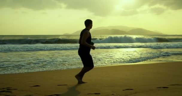 Silhueta de um jovem caucasiano exercitando ao ar livre no fundo de um belo mar.Um homem corre na areia na praia ao pôr do sol contra o pano de fundo do belo mar, grandes ondas bonitas. — Vídeo de Stock