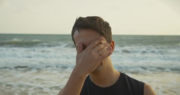 Retrato de um jovem caucasiano olhando para a câmera, ao longo do pano de fundo do belo mar. Praia ao pôr do sol contra o pano de fundo do belo mar, grandes ondas bonitas. — Vídeo de Stock