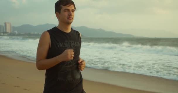 Νεαρός καυκάσιος άνδρας γυμνάζεται με υπαίθριο τρέξιμο με ένα όμορφο φόντο στη θάλασσα. Jogging στην άμμο στην παραλία στο ηλιοβασίλεμα με φόντο την όμορφη θάλασσα, μεγάλα όμορφα κύματα. — Αρχείο Βίντεο