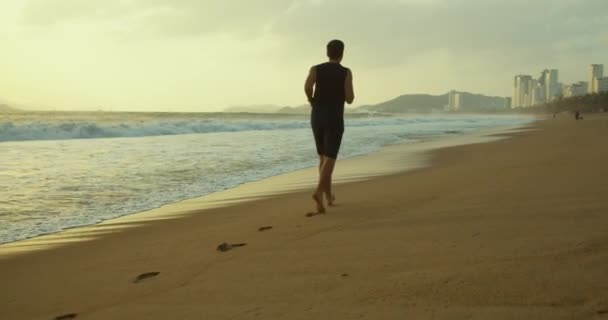 Giovane caucasico uomo si esercitano con la corsa all'aperto con un bellissimo fondo marino. Jogging sulla sabbia sulla spiaggia al tramonto sullo sfondo del bellissimo mare, grandi onde bellissime. — Video Stock