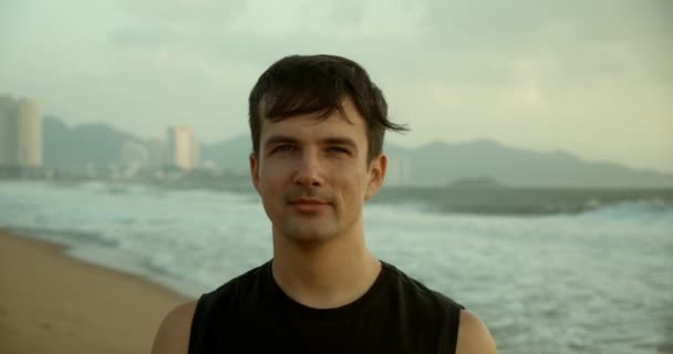 Портрет молодого кавказца, смотрящего в камеру, на фоне прекрасного моря. Пляж на закате на фоне прекрасного моря, больших красивых волн. — стоковое видео