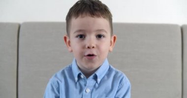 Komik küçük bir çocuğun portresi, gülümseyen bir çocuk, kameraya bakıyor, kanepede oturuyor, güzel yüzlü şirin bir anaokulu çocuğu, şaşırmış bir yüz yapıyor, diyor ki:.