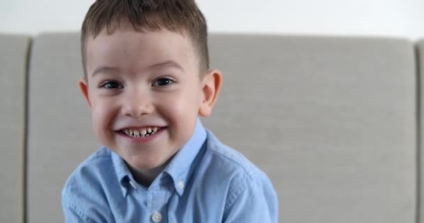 Retrato Engraçado menino sorrindo chil olhando para a câmera estão sentados no sofá em casa, criança pré-escolar bonito com rosto bonito sentado no sofá. Conceito Infância feliz. — Vídeo de Stock