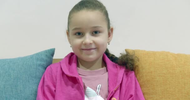 Portrét šťastný legrační holčička usmívající se dítě při pohledu na kameru sedí na gauči doma, roztomilé dítě s hezkou tváří sedí na pohovce. — Stock video