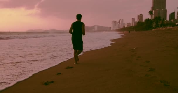 Giovane uomo caucasico che lavora all'aperto sullo sfondo di viola arancio tramonto e bel mare. Correre sulla sabbia sulla spiaggia al tramonto sullo sfondo del bellissimo mare, grandi onde. — Video Stock