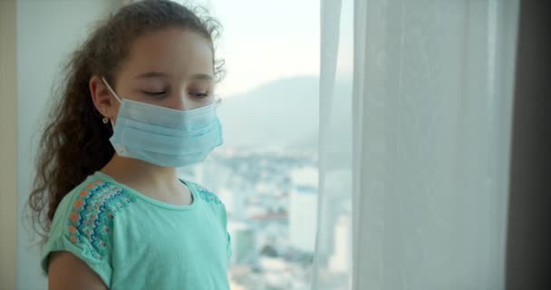 Uma criança com uma máscara médica protetora chega à janela e olha para a rua com um rosto triste e olhos. Pandemia, covid - 19. — Vídeo de Stock